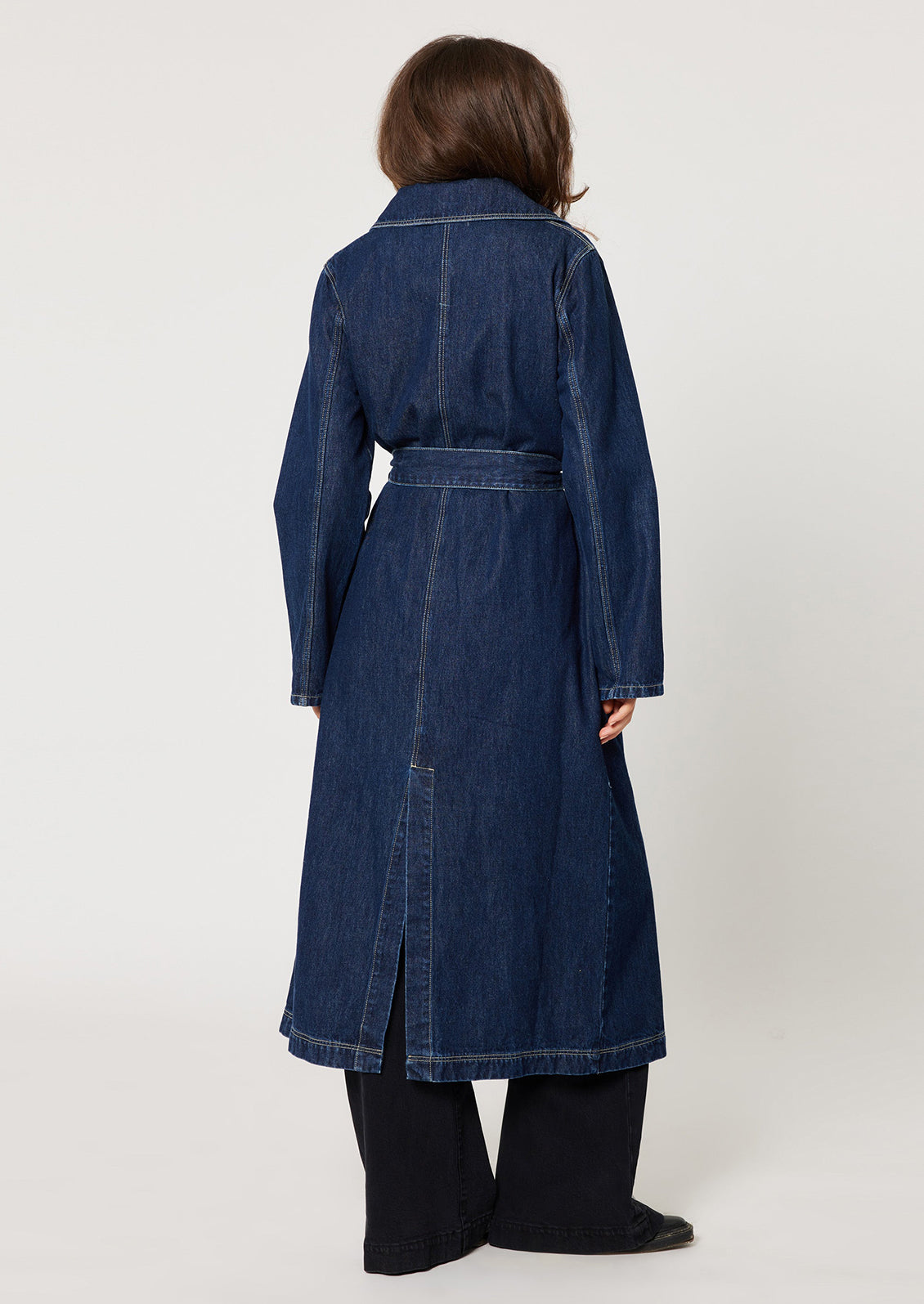 A woman wearing a long denim trench coat in dark blue rinse denim.