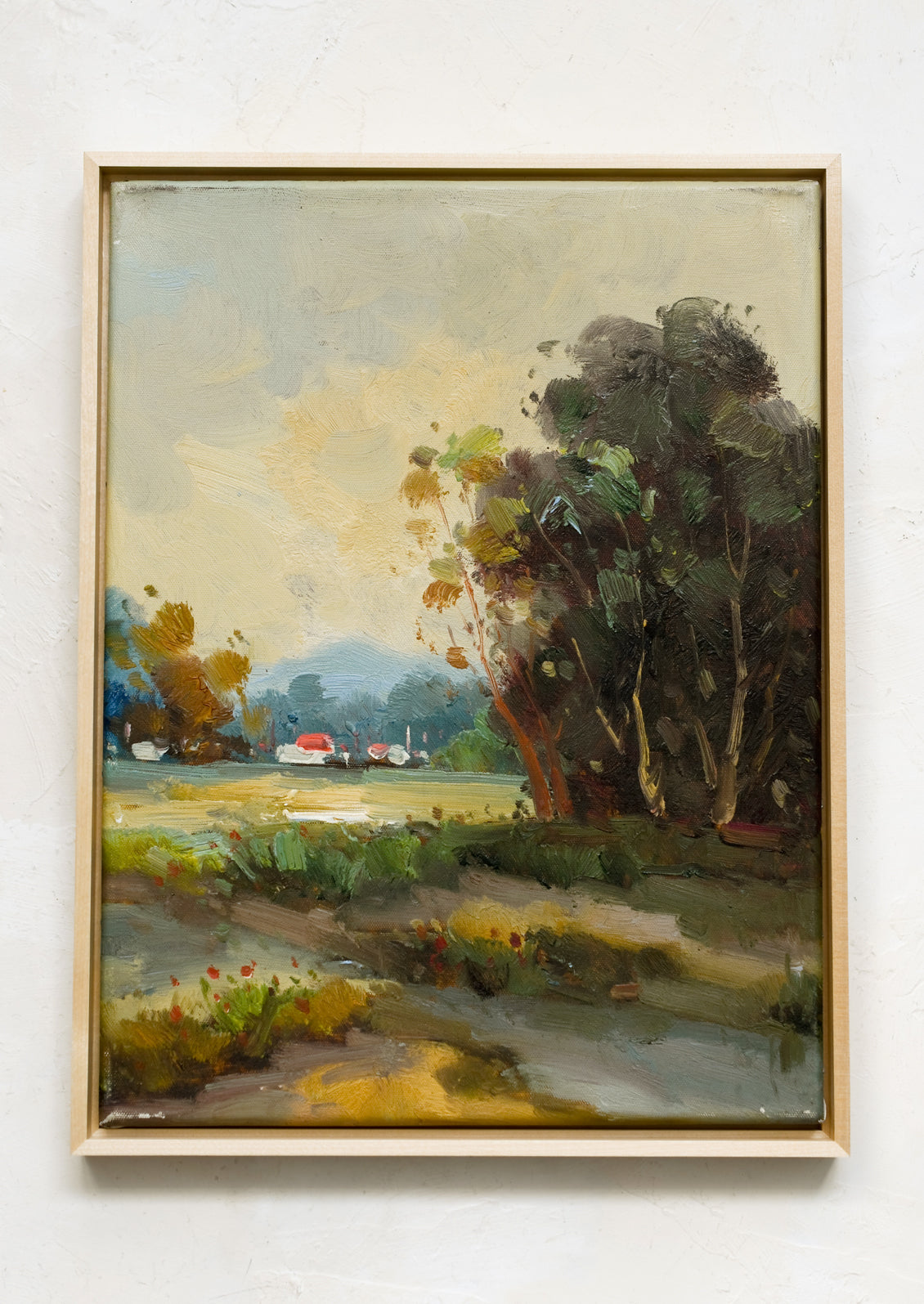A framed landscape oil painting of trees and houses near a creek.