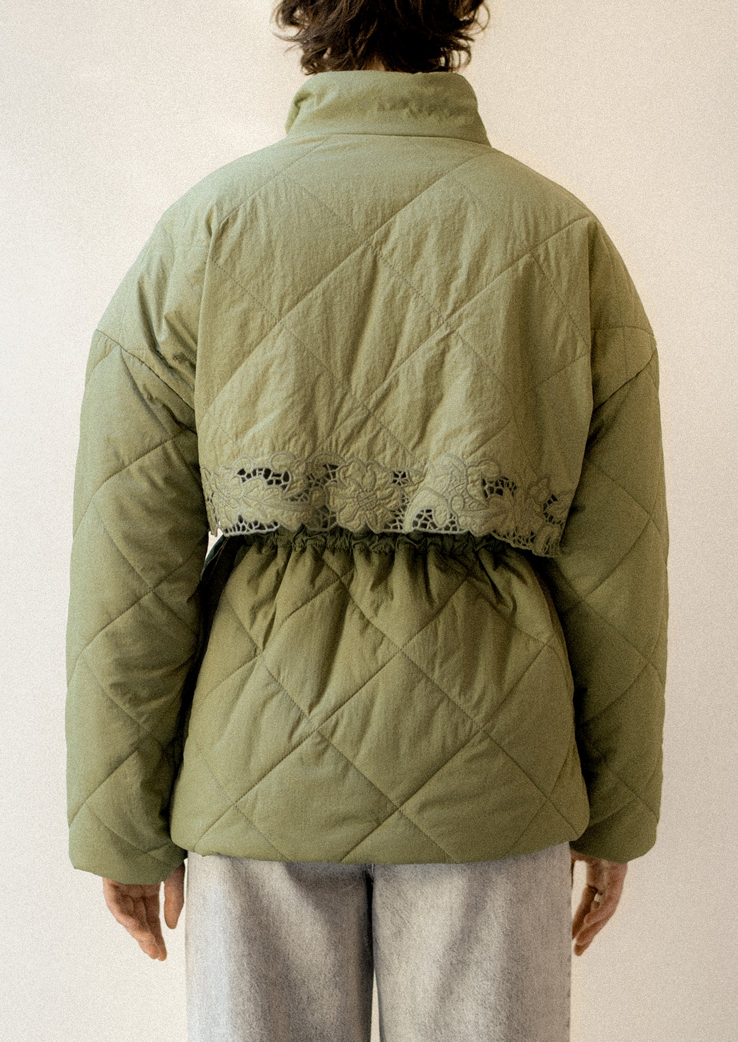 A woman wearing an olive green diamond quilted jacket with lace detailing and drawcord waist.