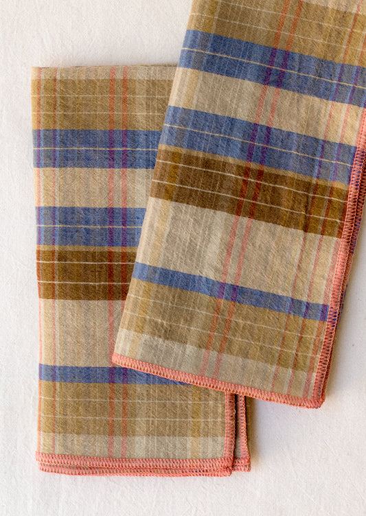 A pair of linen napkins in brown and blue plaid with pink stitch trim.