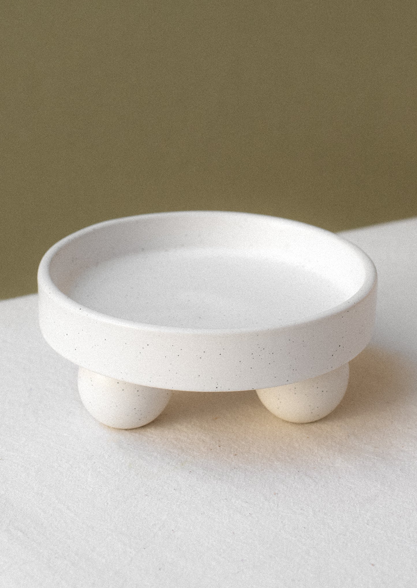 A round, rimmed riser in speckled white ceramic with ball shaped feet.