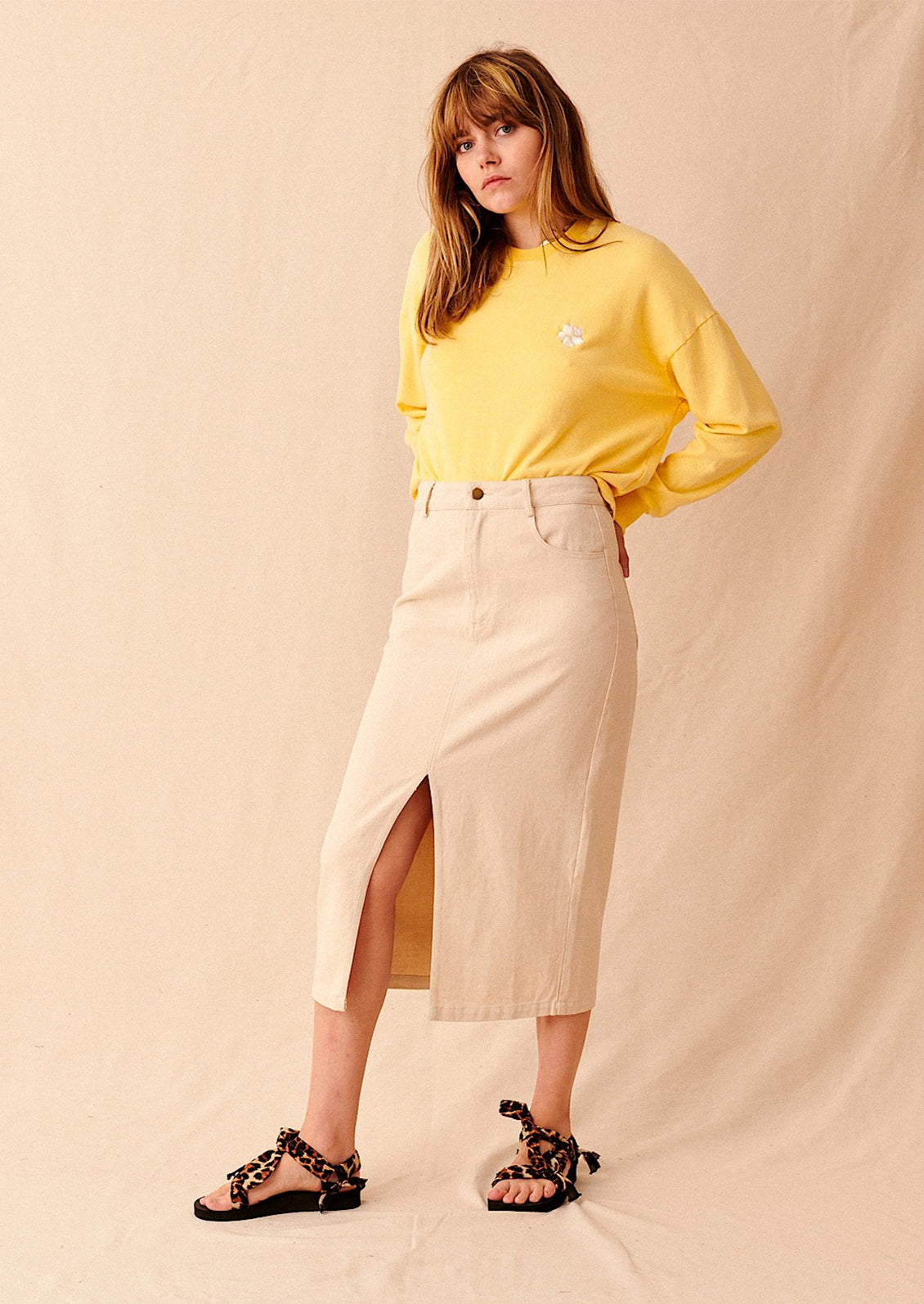 A woman standing with a yellow sweatshirt and a midi length ecru denim skirt