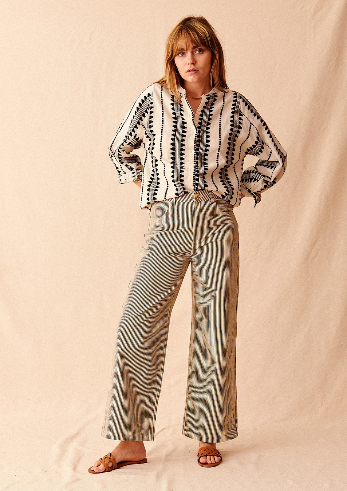 A woman standing wearing cream and black pinstriped wide leg jeans with brown sandals and cream and black embroidered top