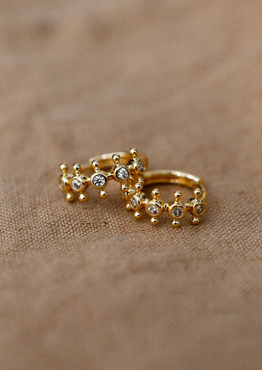 A pair of gold huggie hoop earrings with bezeled clear crystals and decorative ball flange.