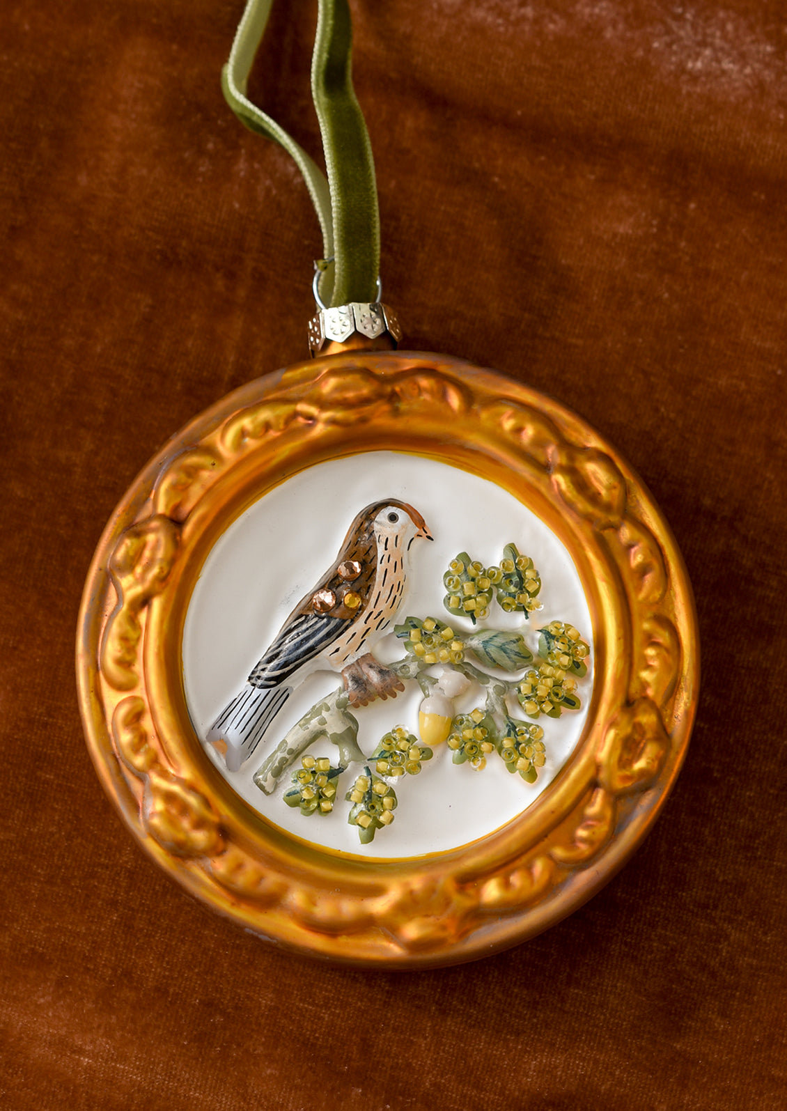 A circular glass ornament with kinglet bird on branch inside gold frame.