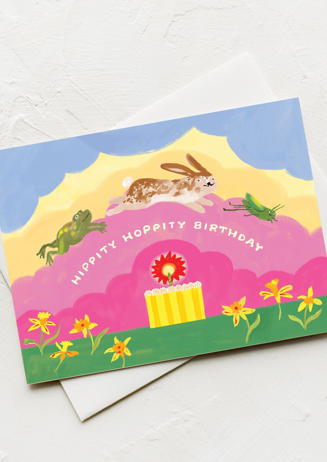 A greeting card with illustration of a frog, rabbit, and cricket jumping over a birthday cake, text reads "Hippity Hoppity Birthday".