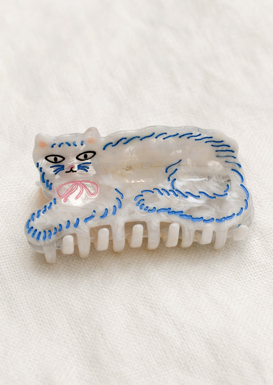 An acrylic hair claw depicting a white cat with blue 'fluff', wearing pink bow collar.