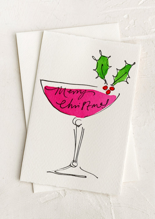 A greeting card with hand painted illustration of a coupe glass decorated with holly leaf, text reads "Merry Christmas".