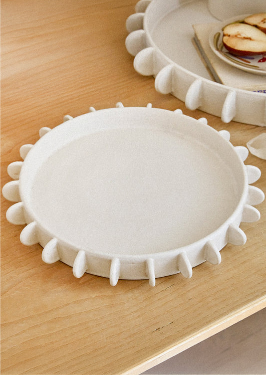 A round shallow ceramic platter in white with decorative tab detailing around outer edge.