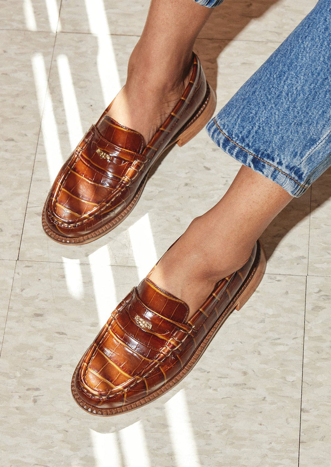 A person wearing a pair of penny loafers in brown croc embossed leather.