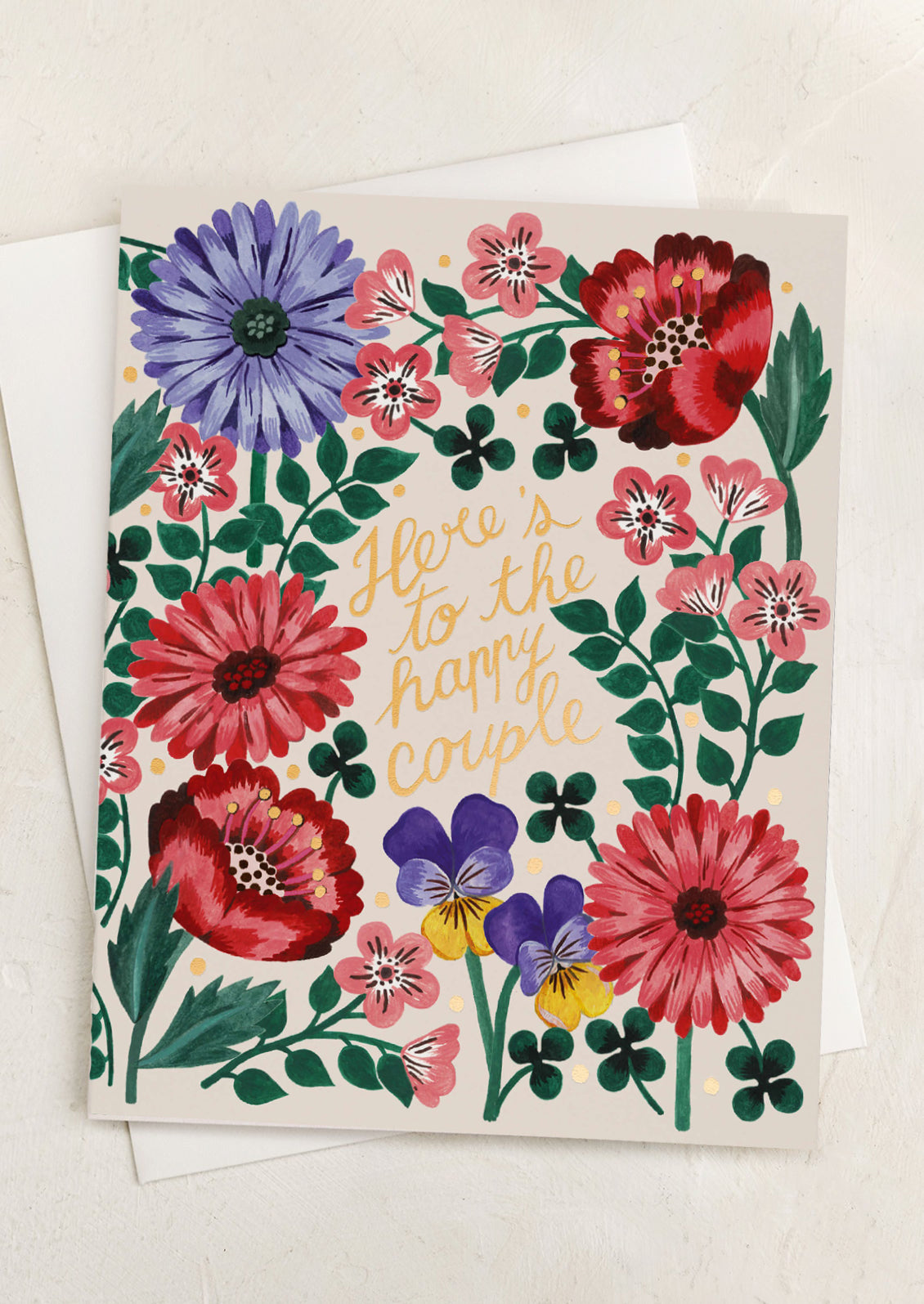 A floral print card, gold text reads "Here's to the happy couple".