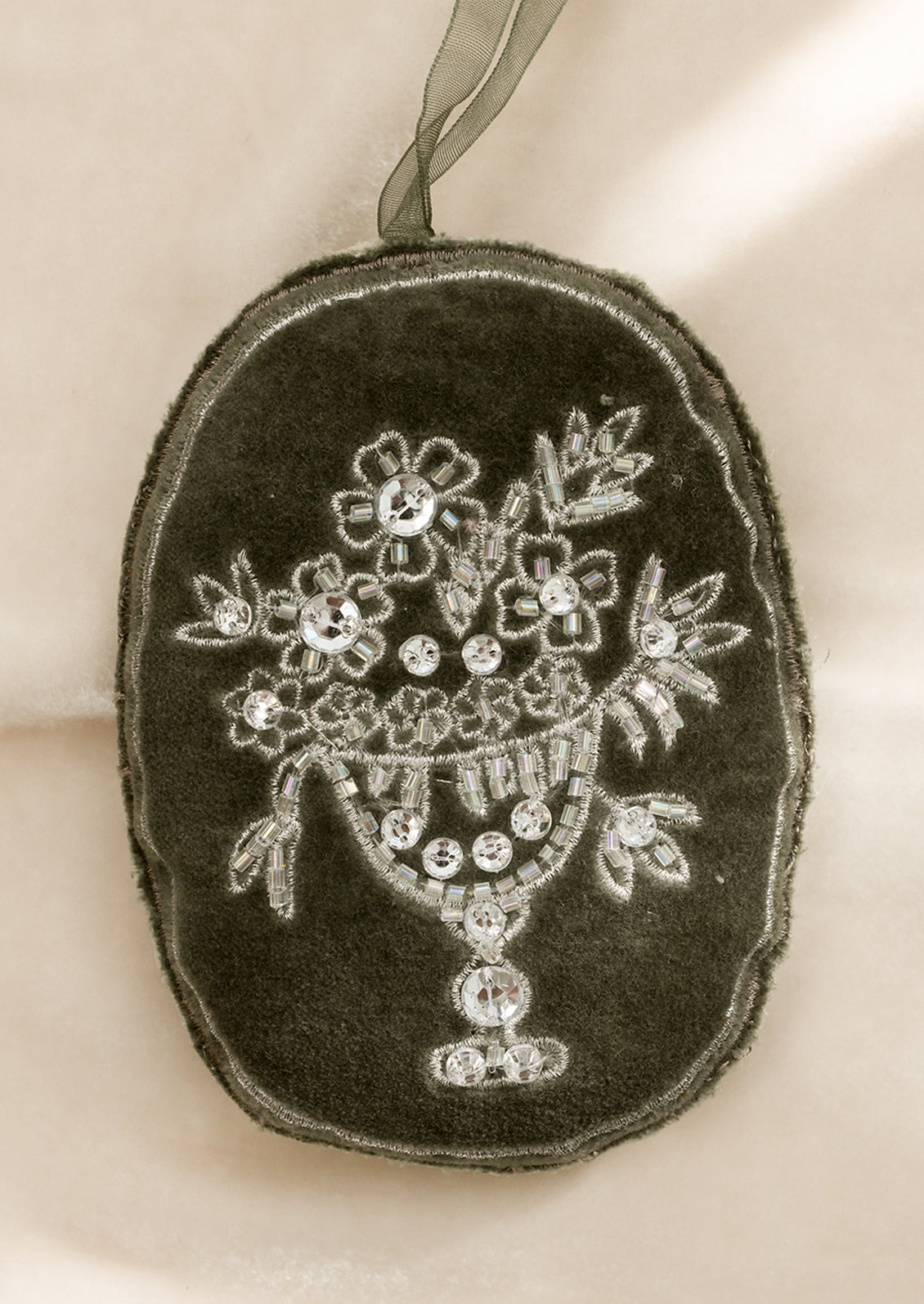 An olive green, oval shaped velvet ornament with silver floral embroidery.