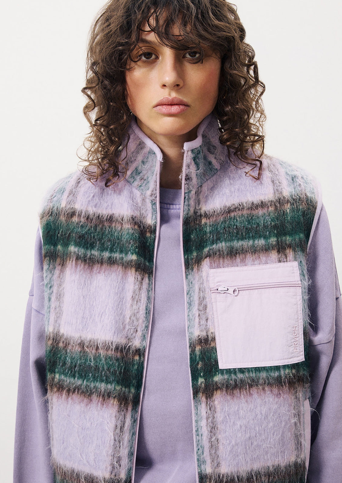 A woman wearing a lavender vest with green plaid pattern and solid zip patch pocket.