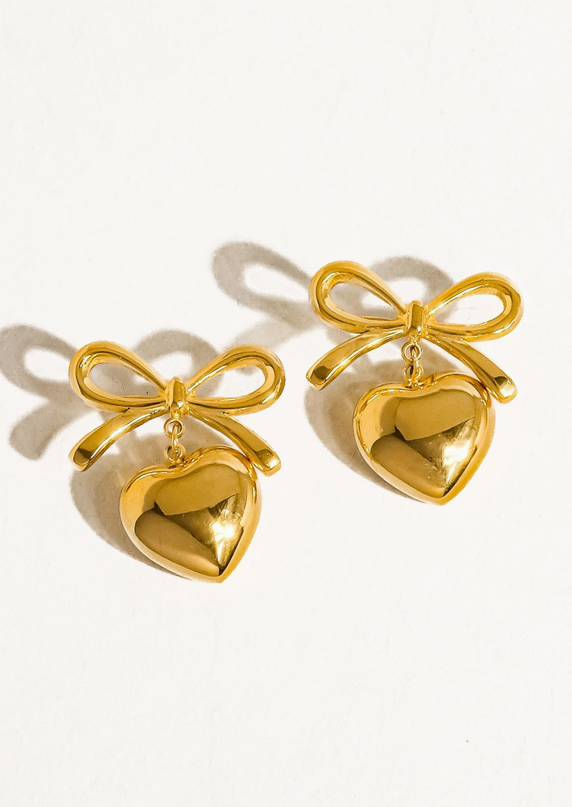 A pair of gold earrings with dangling heart charm and bow shaped post.