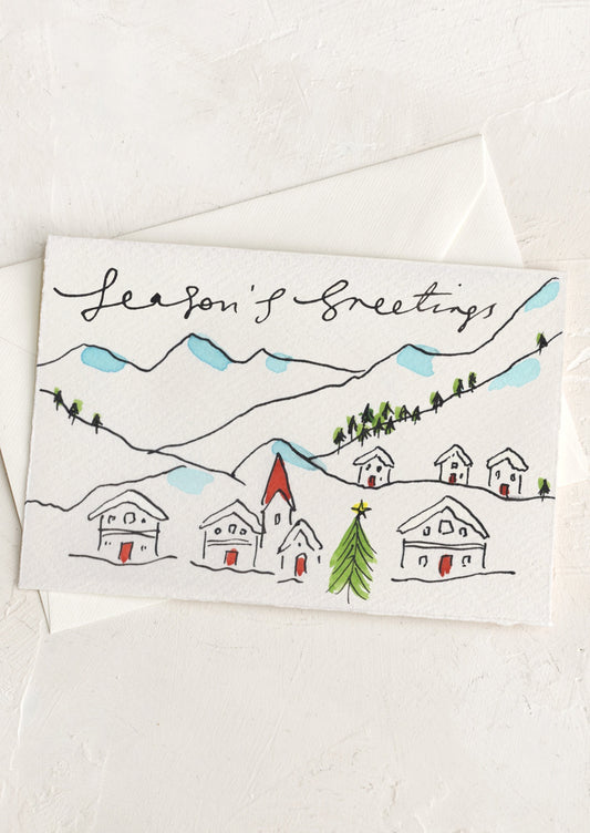 A card with hand painted illustration of holiday homes in the mountains, text reads "Seasons' Greetings".