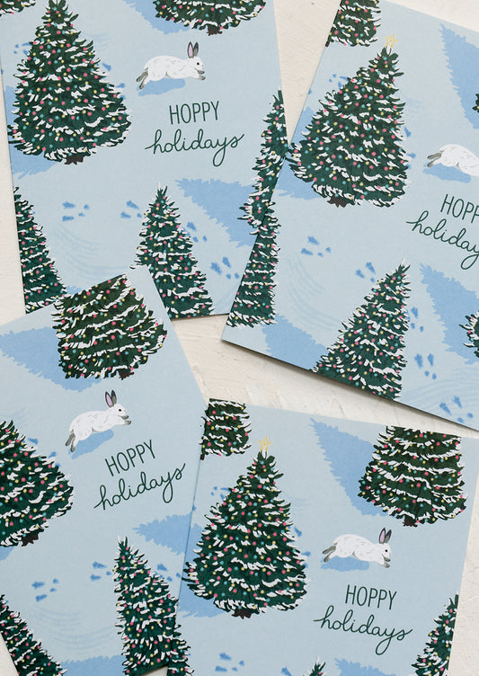 A set of holiday cards with illustration of bunny hopping on snow with Christmas trees, text reads "Hoppy Holidays".