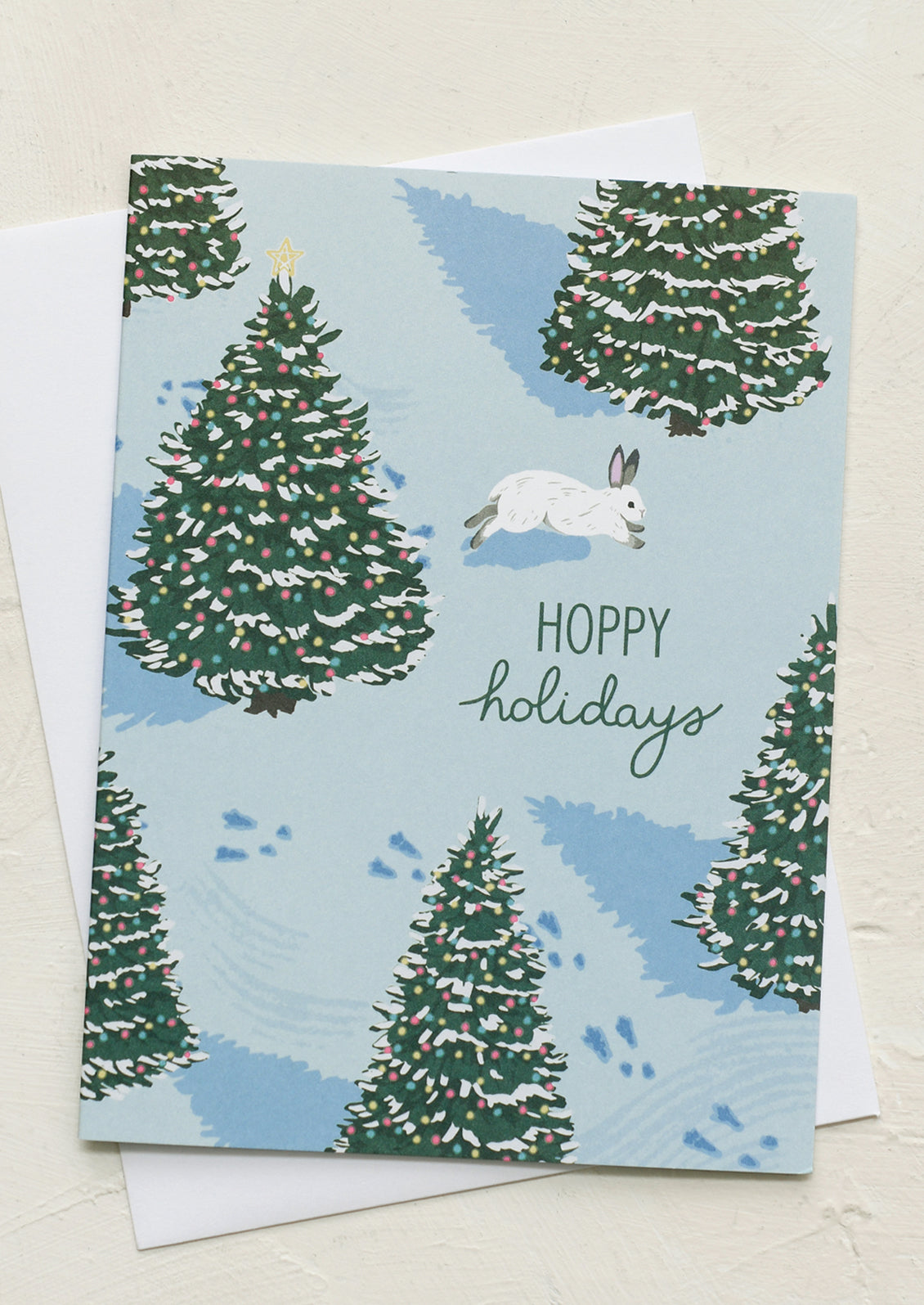 A holiday card with illustration of bunny hopping on snow with Christmas trees, text reads "Hoppy Holidays".