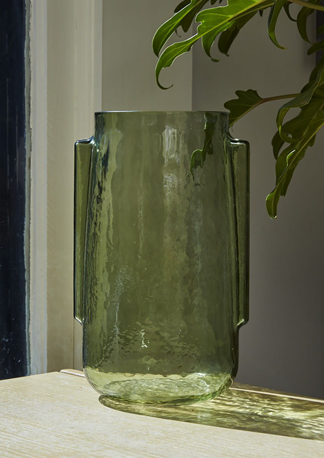 A transparent green glass vase in tall shape with side handle detail.