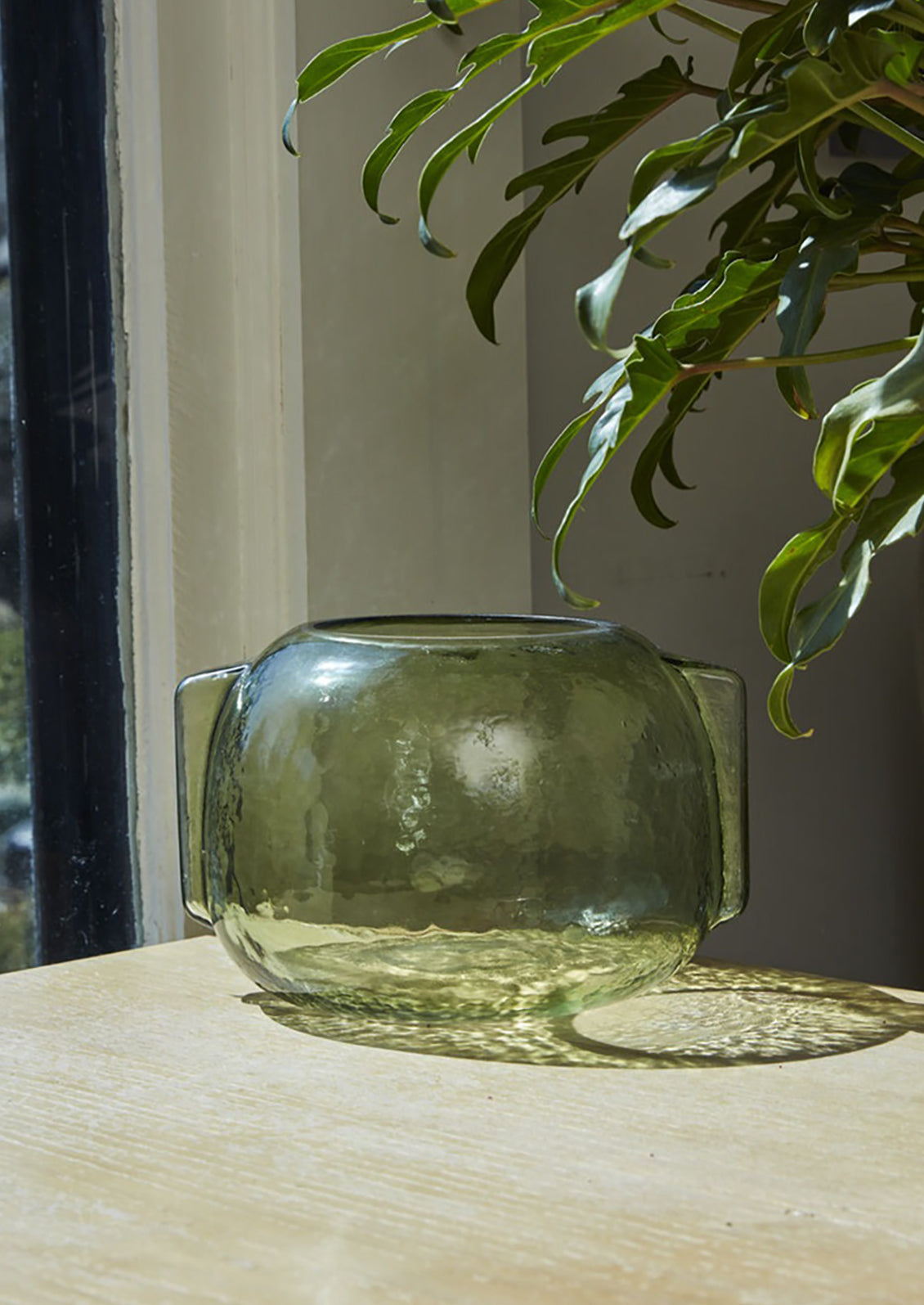 A transparent green glass vase in short/wide shape with side handle detail.