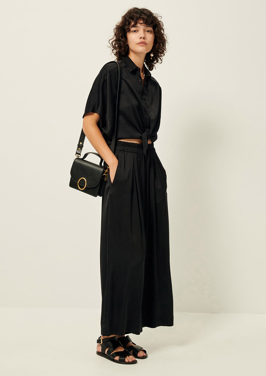 Woman wearing black flowy pants, black button-up, sandals, and purse. 