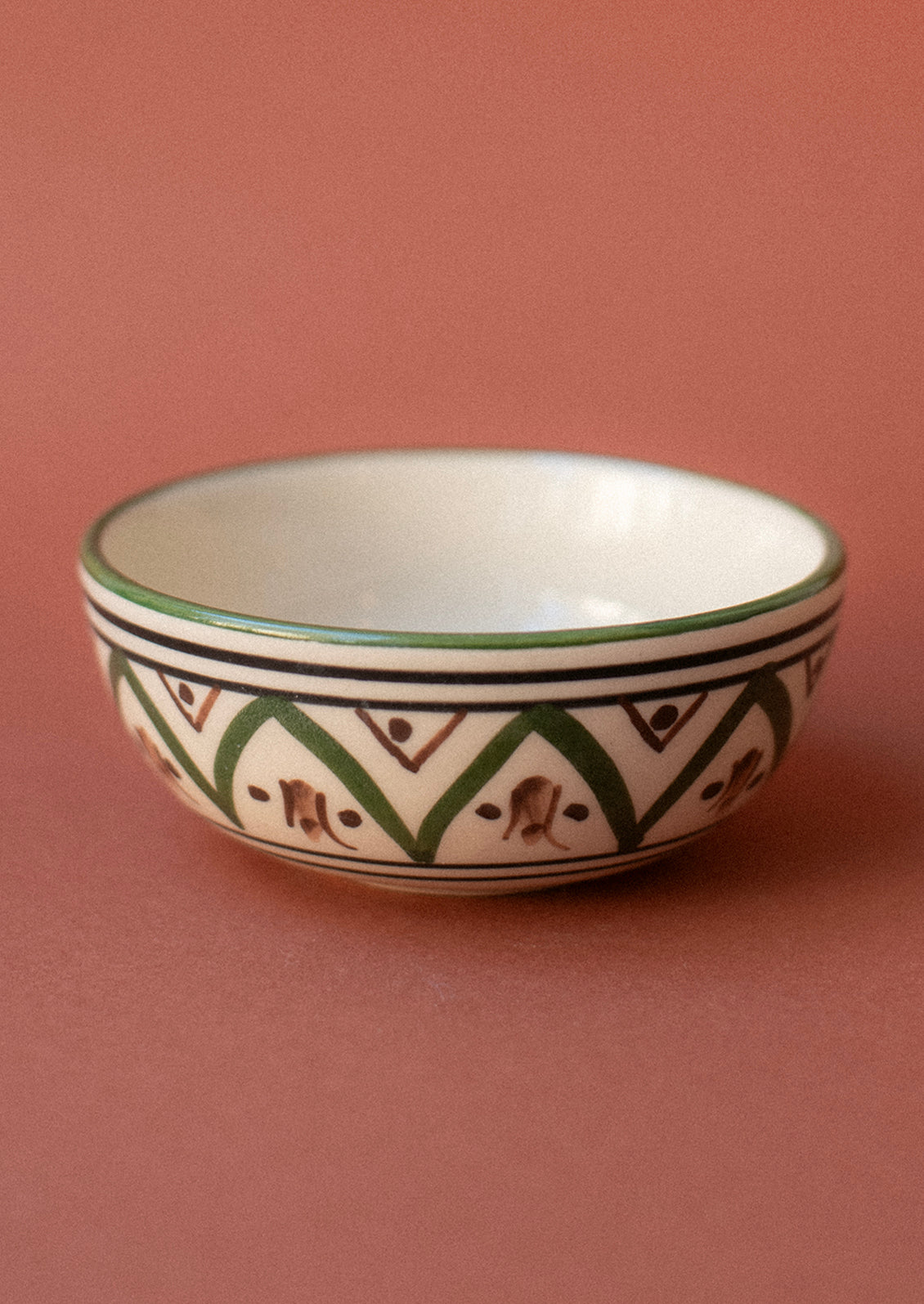 A small ceramic pinch bowl in cream with Moroccan-inspired painted geometric design in green and brown.