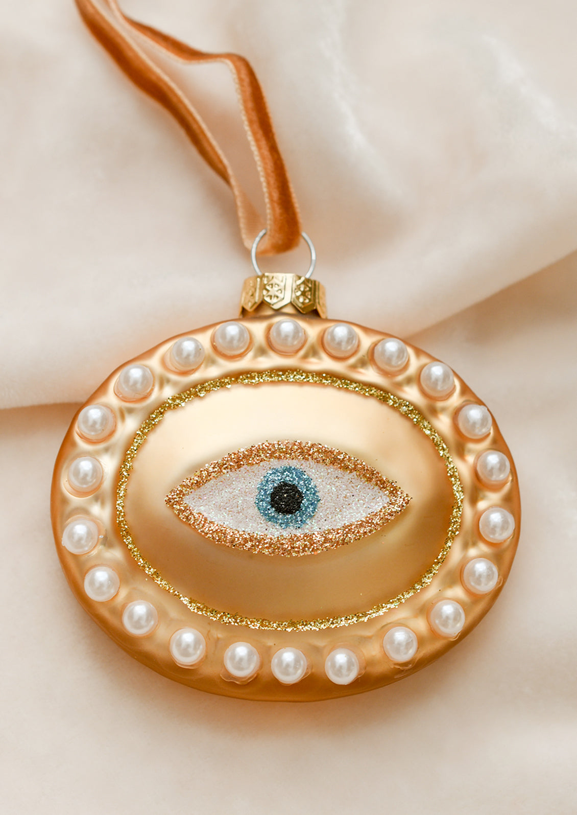 A glass ornament of gold oval with pearl border and glittery eyeball at center.