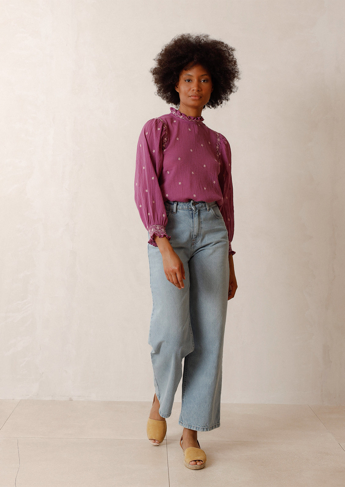 Woman wearing magenta top with embroidered details, blue jeans, and flats. 