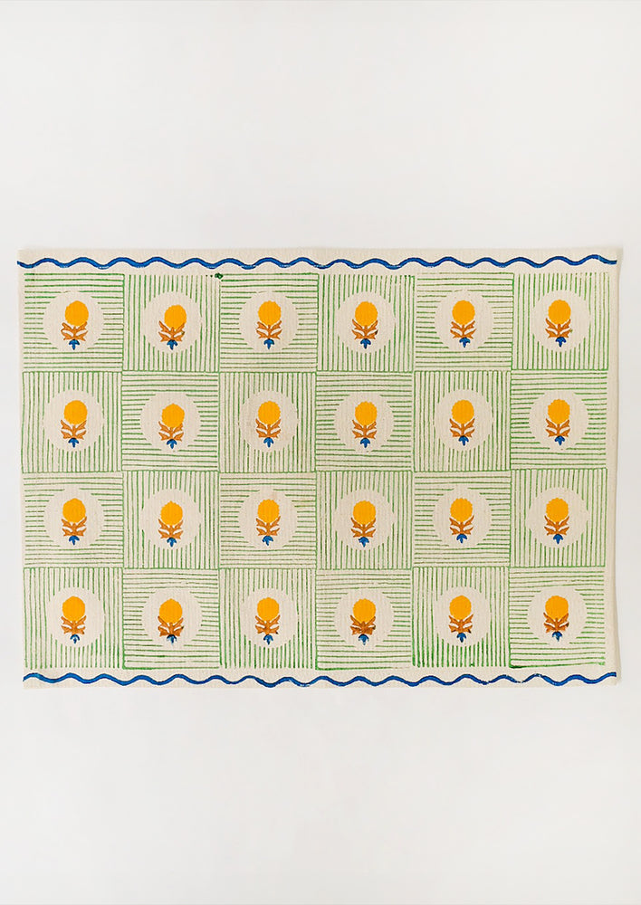 A block printed placemat in natural linen with green squares, yellow floral and blue wavy trim.