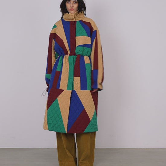 A woman wearing a geometric patchwork quilted coat in wine, teal, beige and blue.