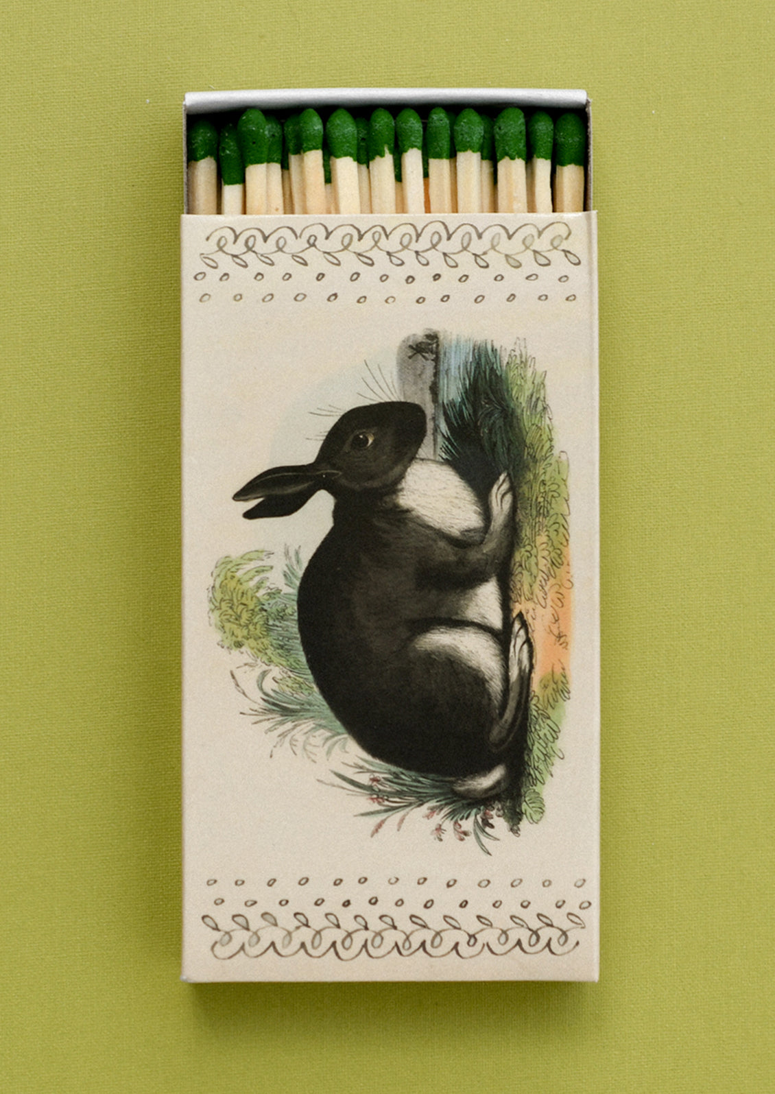 A matchbox with imagery of black rabbit, holding green tipped matches.