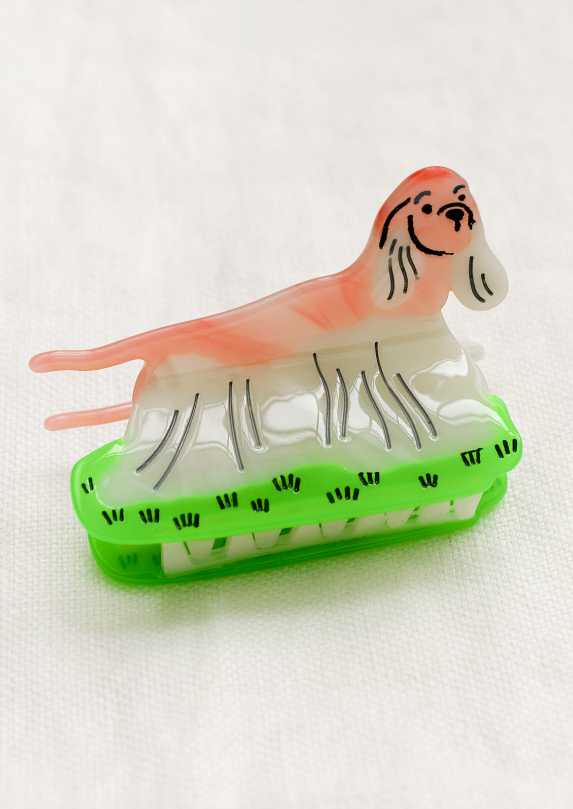 A hair claw in the shape of a spaniel breed dog in pink and white, standing on neon green grass patch.