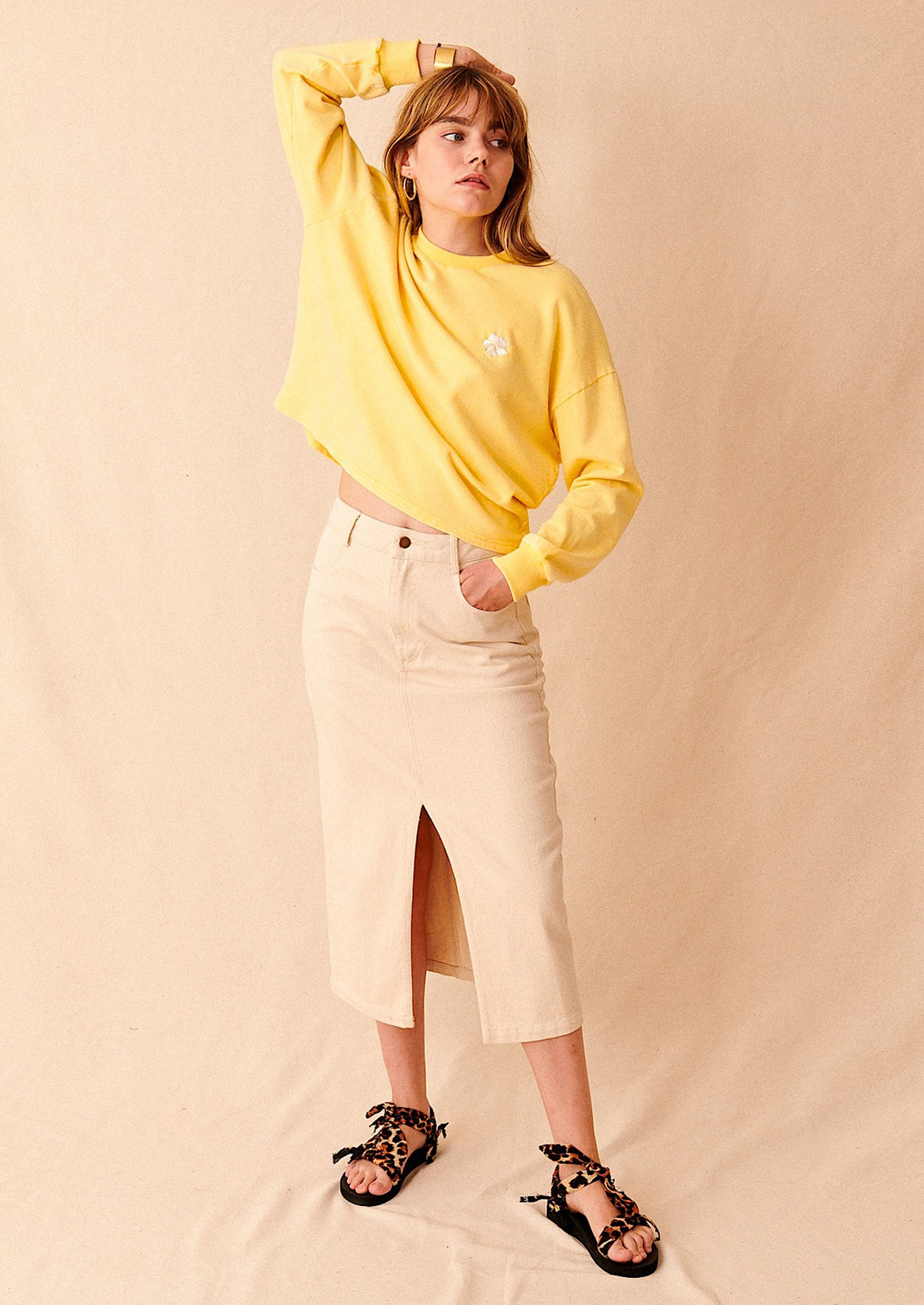 A woman standing with a yellow sweatshirt and a midi length ecru denim skirt
