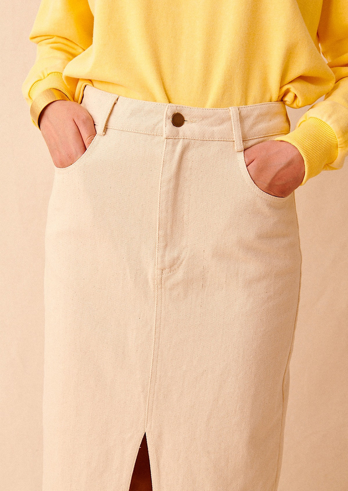 A person wearing a yellow sweatshirt tucked into an ecru denim skirt with their hands in pockets