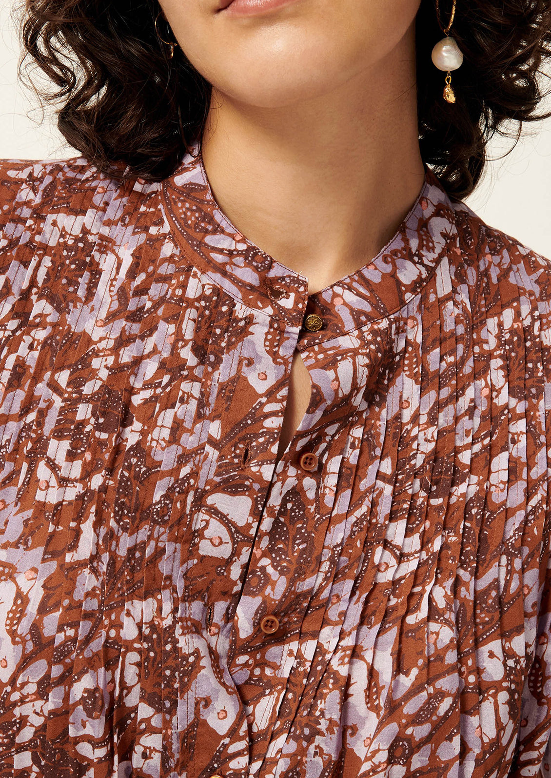 A woman wearing an abstract printed purple and rust colored button up top, showing pintucking and collar detail.