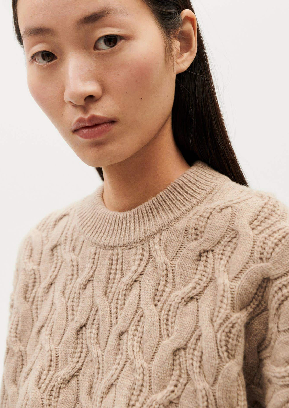 A woman wearing a tan crewneck sweater with braided texture.