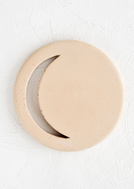 A round concrete trivet in straw color with crescent moon shaped cutout.