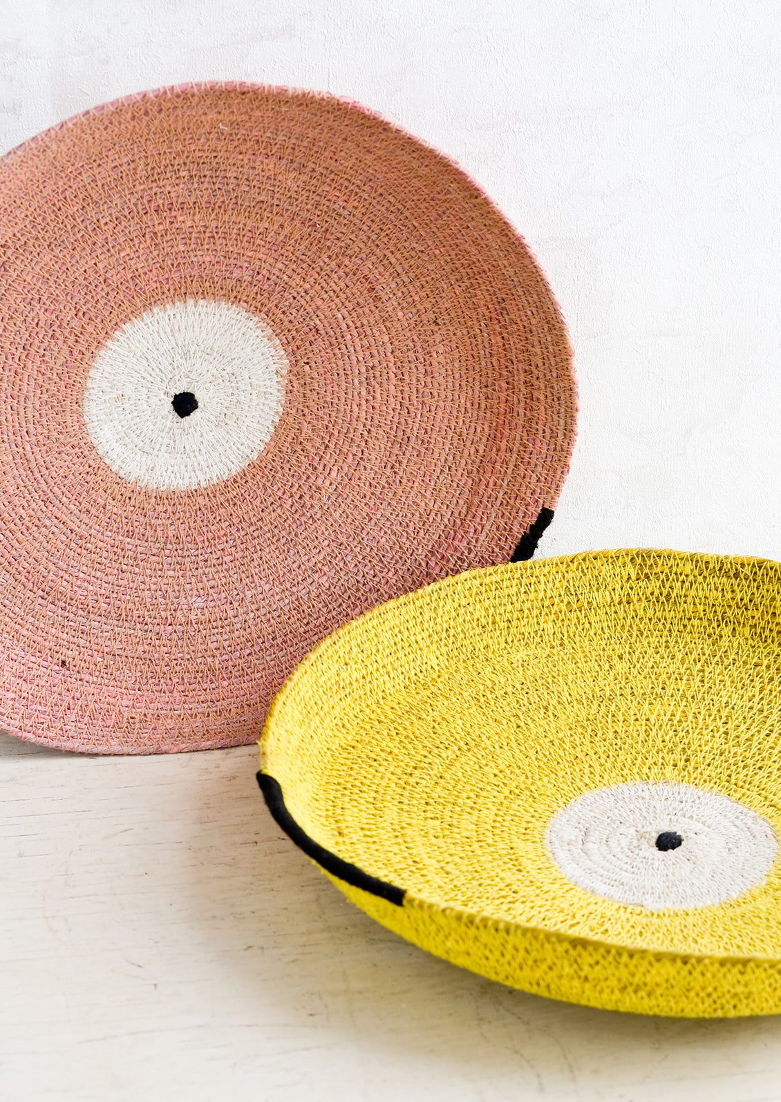 Two round platters/trays made from woven seagrass in yellow and pink.