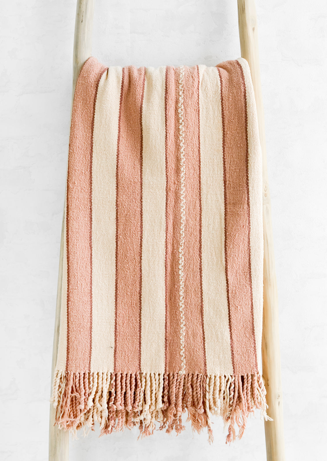 Natural woven cotton throw in blush and cream stripes with twisted fringe edge, hung on a display ladder.