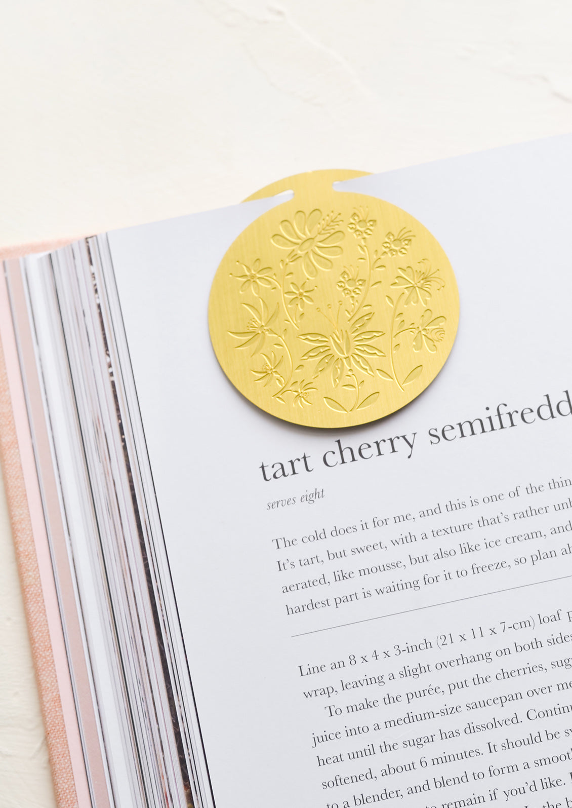 A circular brass bookmark with etched botanical pattern, marking a recipe page.