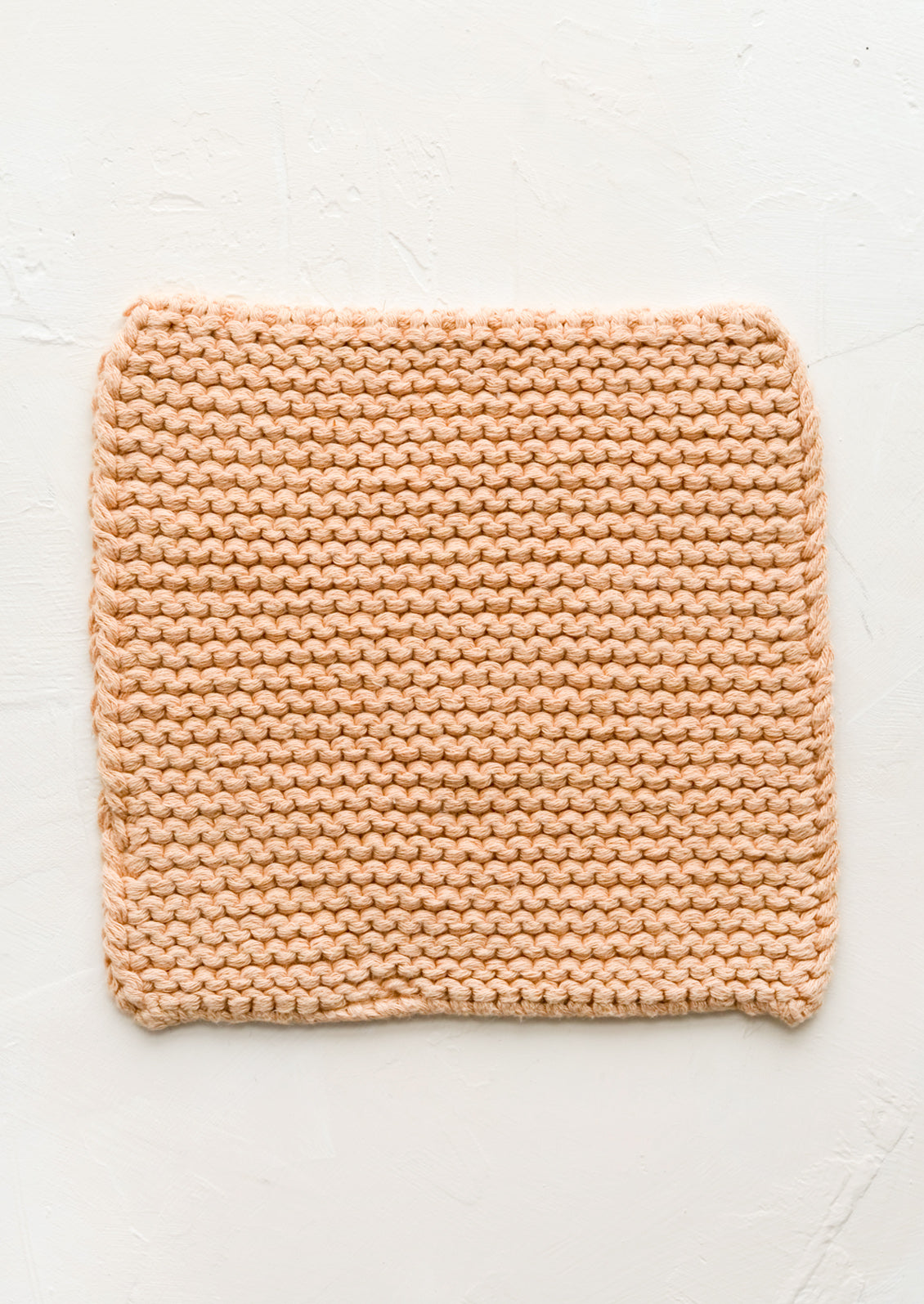 A square, chunky knit cotton potholder in soft peach color.