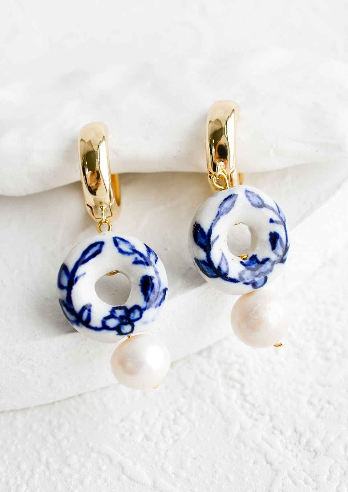 A pair of gold huggie hoops with donut shaped blue and white ceramic bead and pearl below.