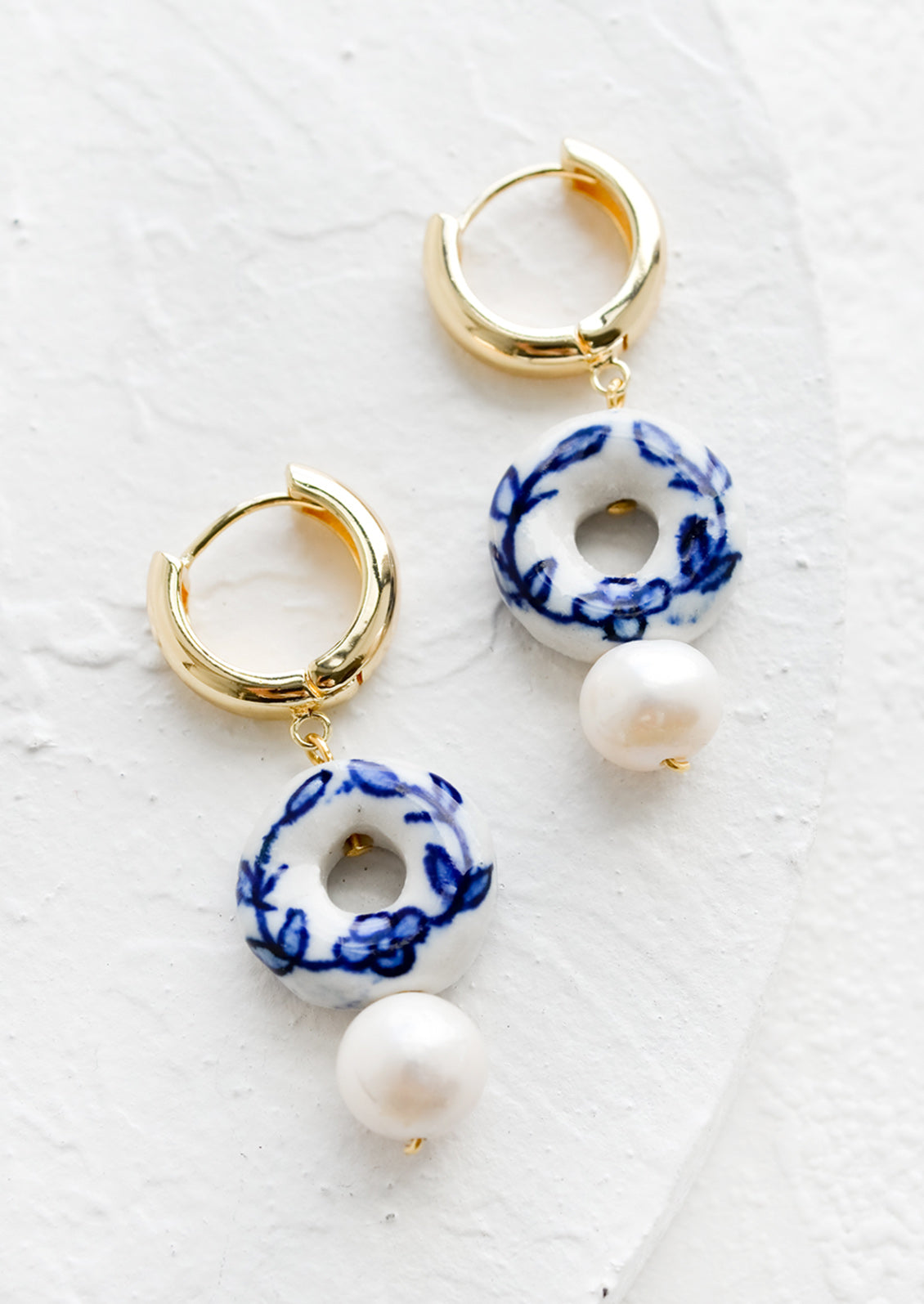 A pair of gold huggie hoops with donut shaped blue and white ceramic bead and pearl below.