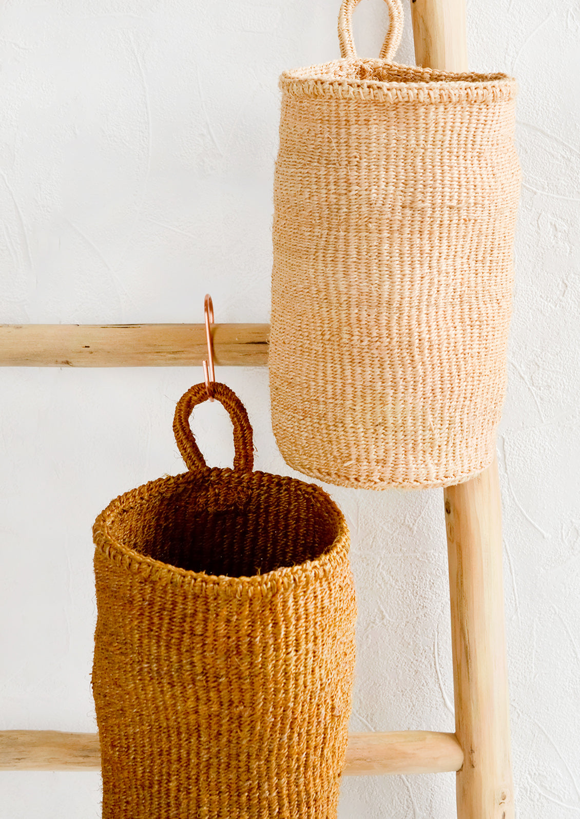 Two oblong cylindrical hanging baskets made from woven sisal, hanging on a display ladder.