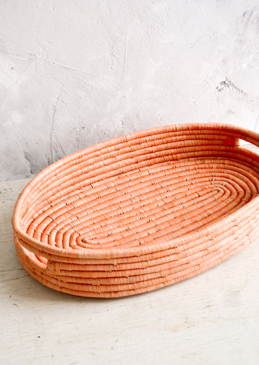 Oval shaped, shallow basket woven in peach raffia with cutout handles at sides.