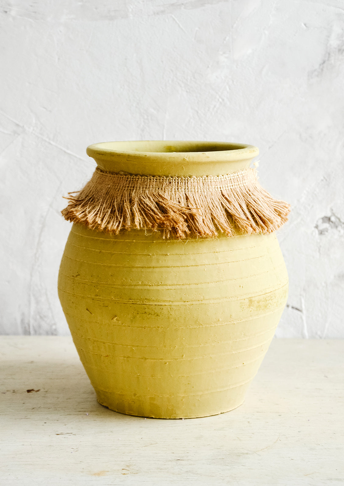 Tall, ochre colored clay vase with fringed jute trim around opening