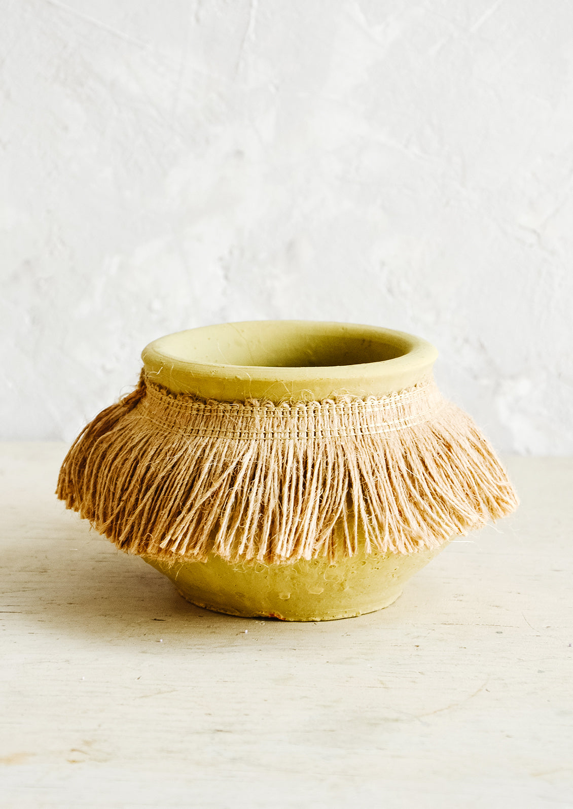 Short, ochre colored clay vase with fringed jute trim around opening