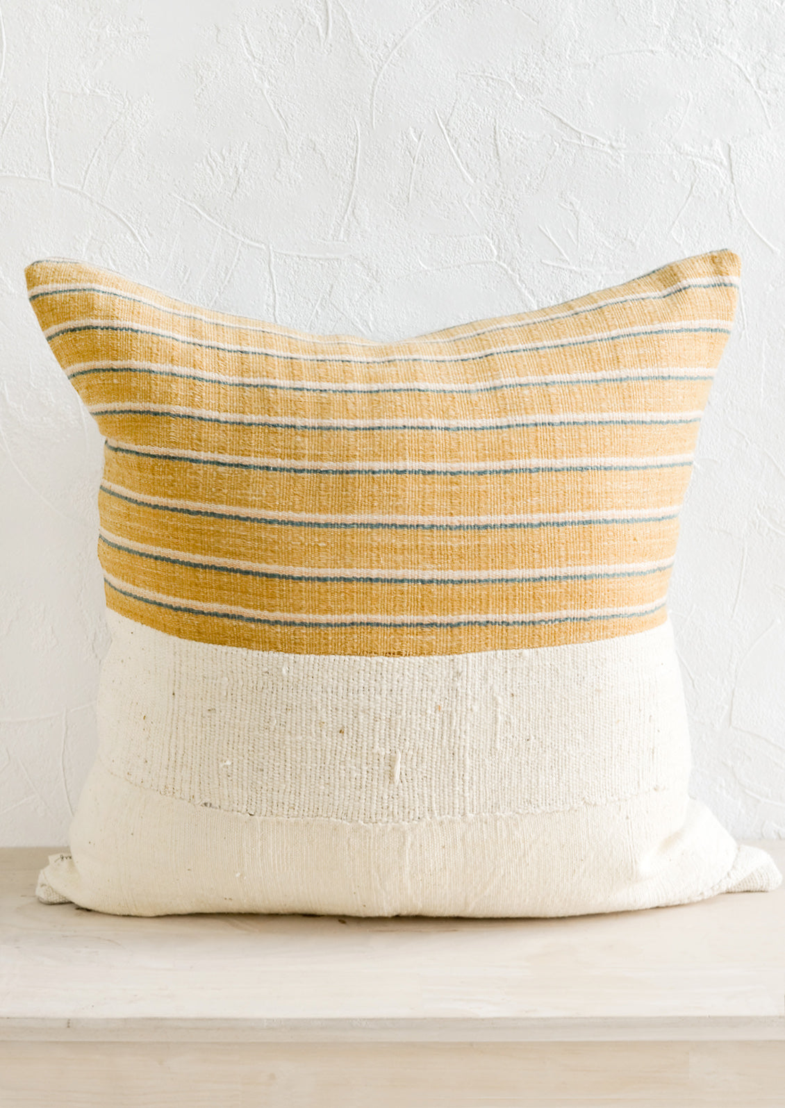 A throw pillow with top half in mustard, ivory & teal striped fabric and bottom half in natural mudcloth.