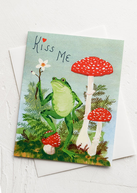 A greeting card with illustration of frog standing on toadstools, text reads "Kiss me".