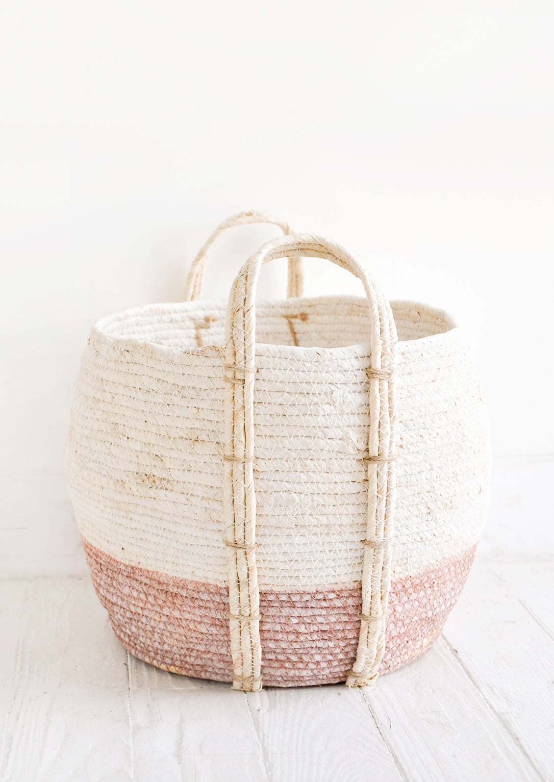 Round storage basket made from natural maize fiber, fiber handles attached at sides, band of contrasting pink color along bottom.
