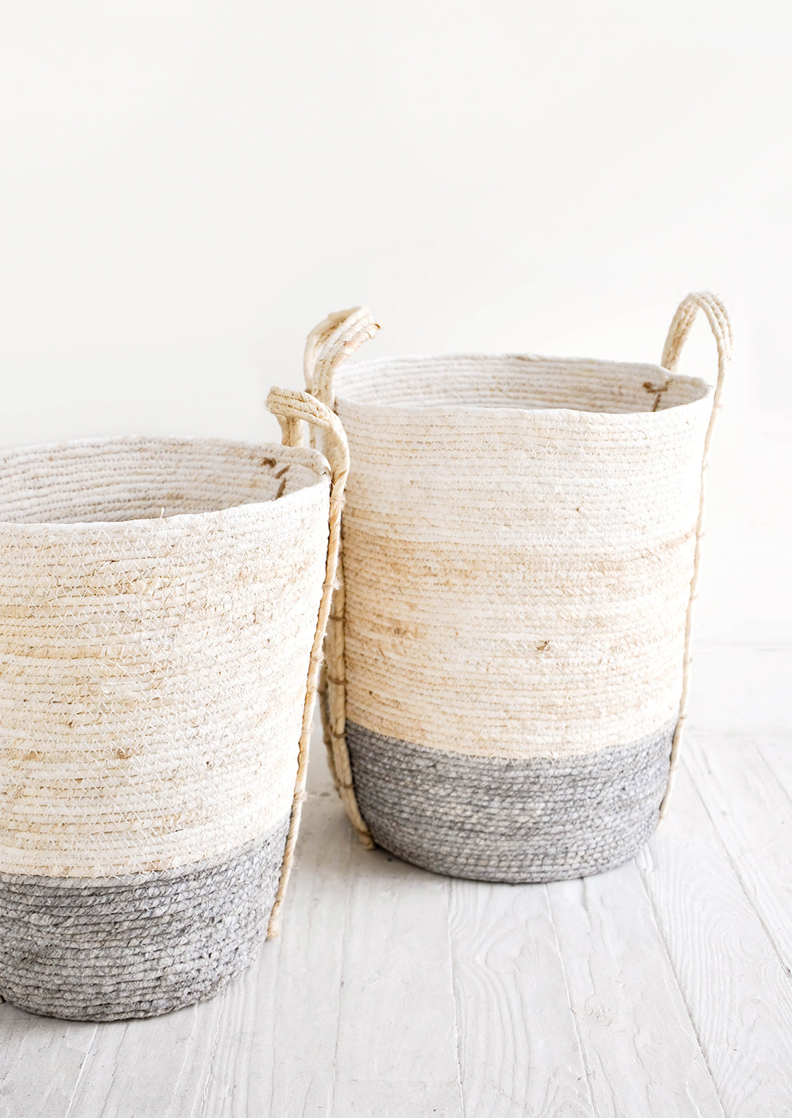 Tall, round storage baskets made from natural maize fiber, fiber handles attached at sides, band of contrasting grey color along bottom.